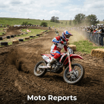 Motocross rider in red and blue gear on number 7 bike turning on dirt track with spectators behind fence.